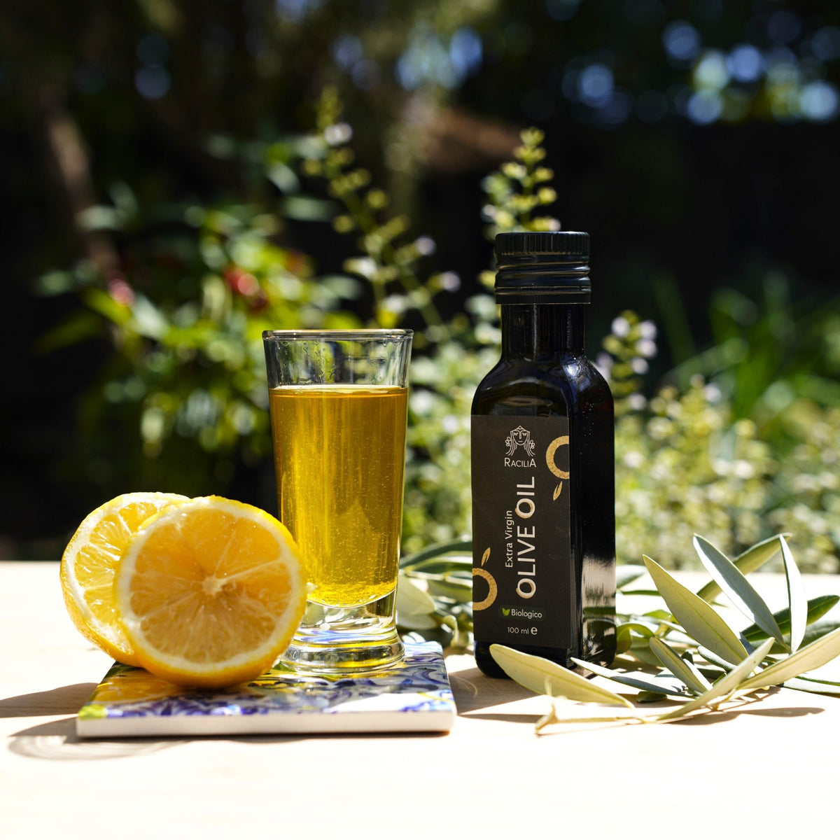 Huile d'olive bio Racilia 100 ml avec un verre d'huile, des citrons et une majolique sicilienne